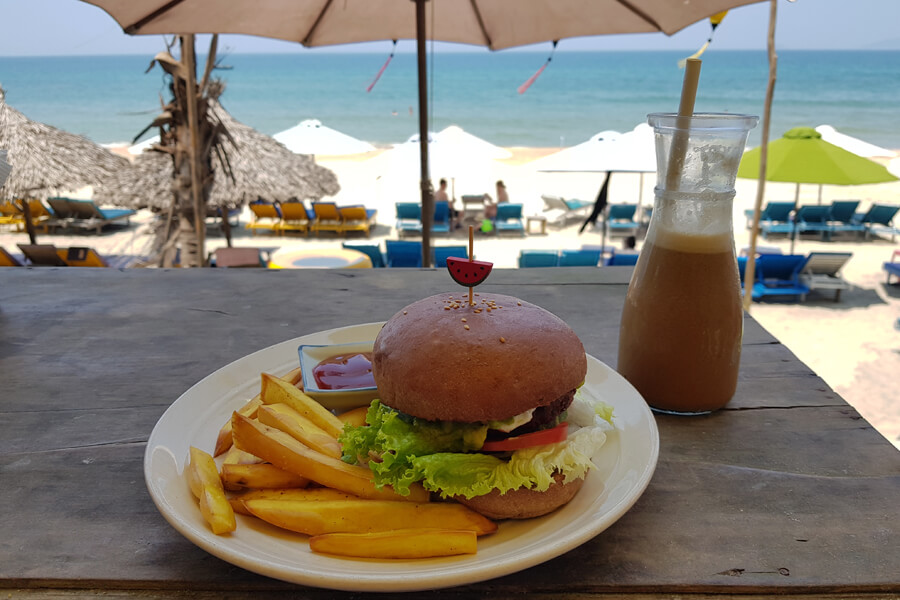 Ein Teller mit Burger und Pommes und ein Eiskaffe stehen auf einem Tisch mit Ausblick auf das Meer.