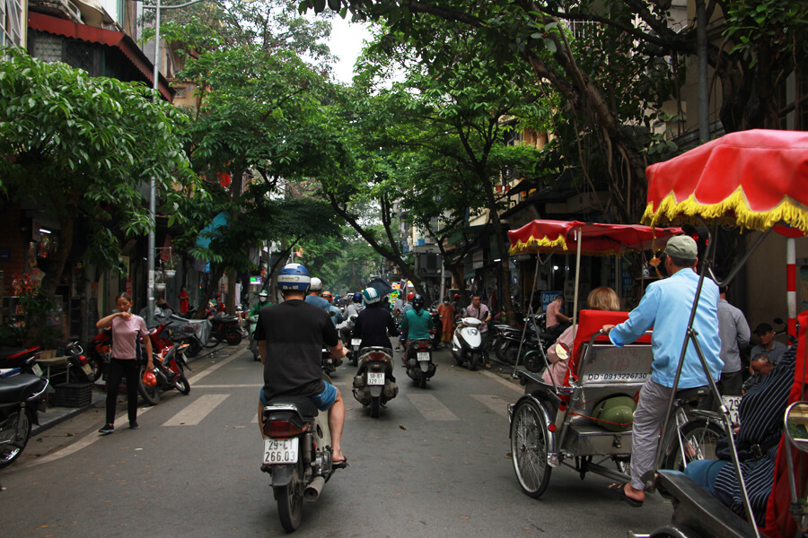 Motorroller und Rikschas fahren über eine Baumgesäumte Straße in der Altstadt von Hanoi