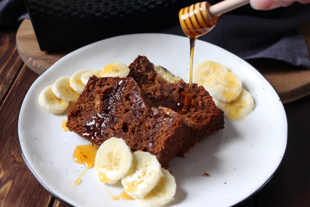 veganes Bananenbrot mit Bananenscheiben, mit einem Honiglöffel wird veganer Honig darüber verteilt