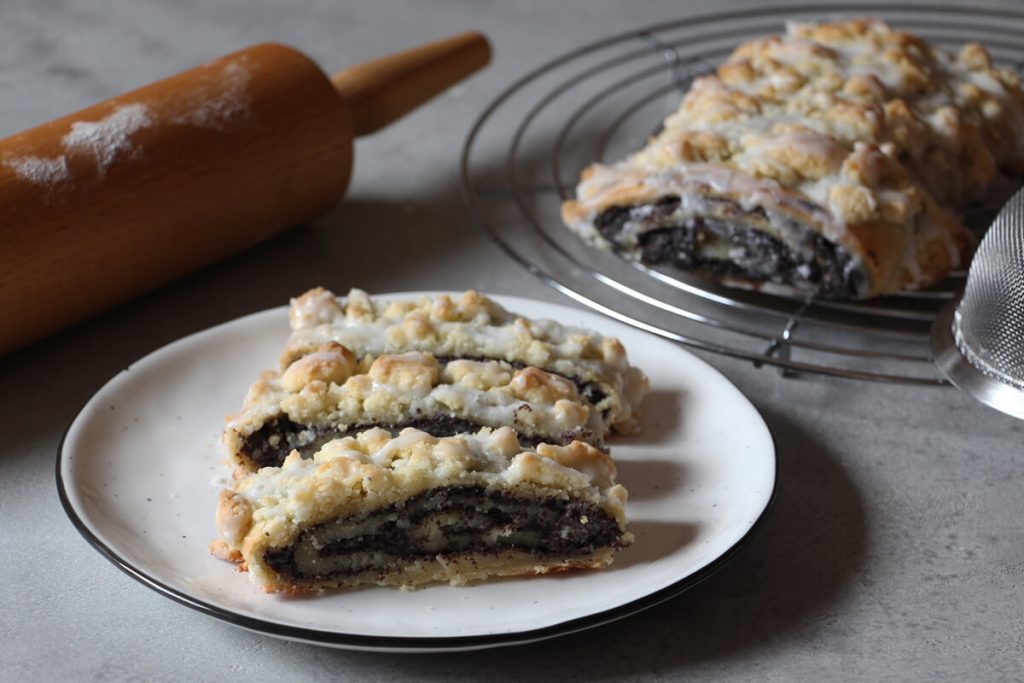 drei Stücke veganer Mohnstriezel auf einem Teller, daneben ein Nudelholz und ein weiterer ganzer Mohnstriezel auf einem Kuchenrost