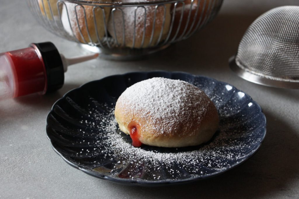 veganer Ofenberliner mit Puderzucker auf einem Teller, daneben eine Spritztülle mit Marmelade, ein Sieb und ein Korb mit weiteren Berlinern