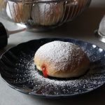 veganer Ofenberliner mit Puderzucker auf einem Teller, daneben eine Spritztülle mit Marmelade, ein Sieb und ein Korb mit weiteren Berlinern