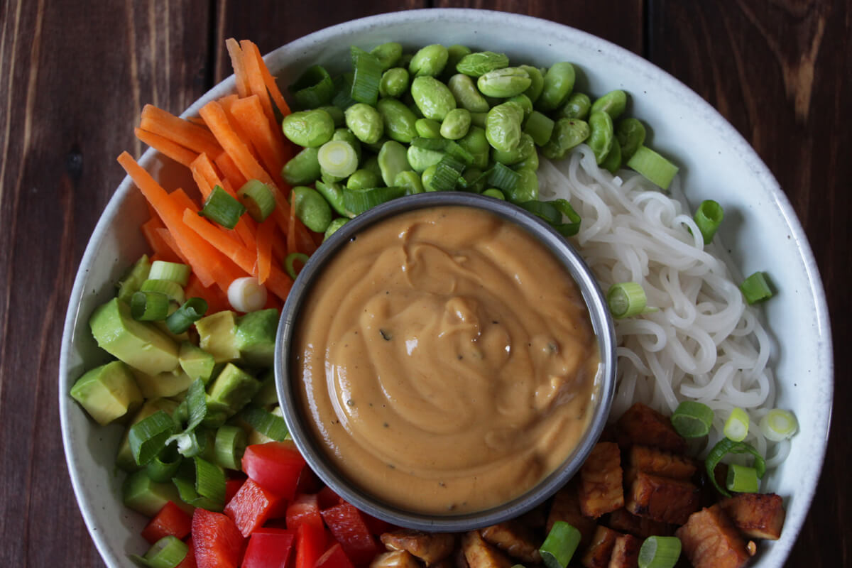 vegane Reisnudelbowl mit frischem Gemüse, angebratenem Tempeh und einer Schale mit Erdnussoße in der Mitte