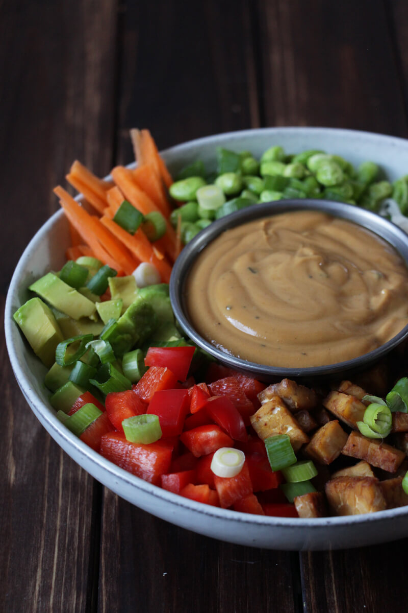vegane Reisnudelbowl mit frischem Gemüse, angebratenem Tempeh und einer Schale mit Erdnussoße in der Mitte