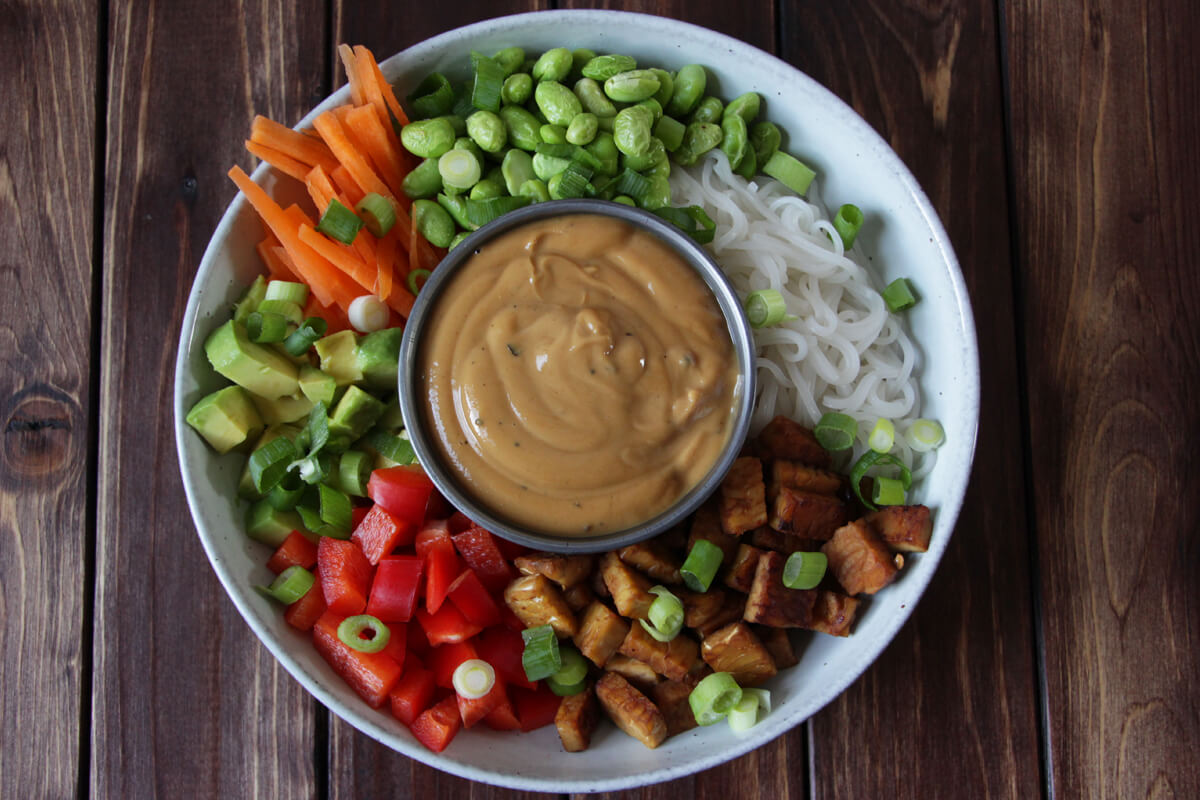 vegane Reisnudelbowl mit frischem Gemüse, angebratenem Tempeh und einer Schale mit Erdnussoße in der Mitte