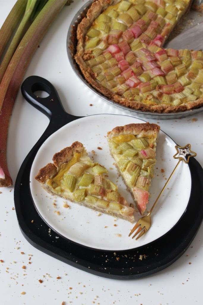 Zwei Stücke vegane Rhabarbertarte mit einer Gabel auf einem Teller, im Hintergrund die restliche Tarte und ein paar Rhabarberstangen.