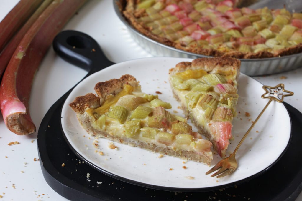 Zwei Stücke vegane Rhabarbertarte mit einer Gabel auf einem Teller, im Hintergrund die restliche Tarte und ein paar Rhabarberstangen.
