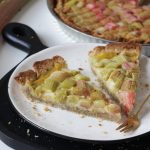 Zwei Stücke vegane Rhabarbertarte mit einer Gabel auf einem Teller, im Hintergrund die restliche Tarte und ein paar Rhabarberstangen.