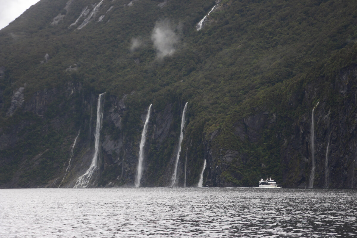 Ein kleines Boot fährt neben hohen Wasserfällen im Milford Sound